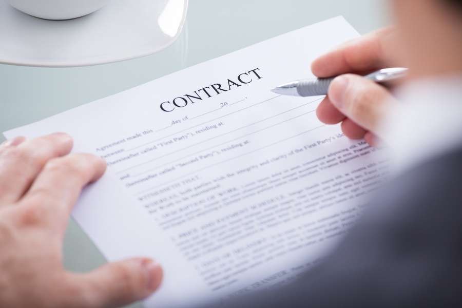 Une personne tient un stylo pour signer un document intitulé "Contract", illustrant la concrétisation de l'achat d'un immeuble de rapport.