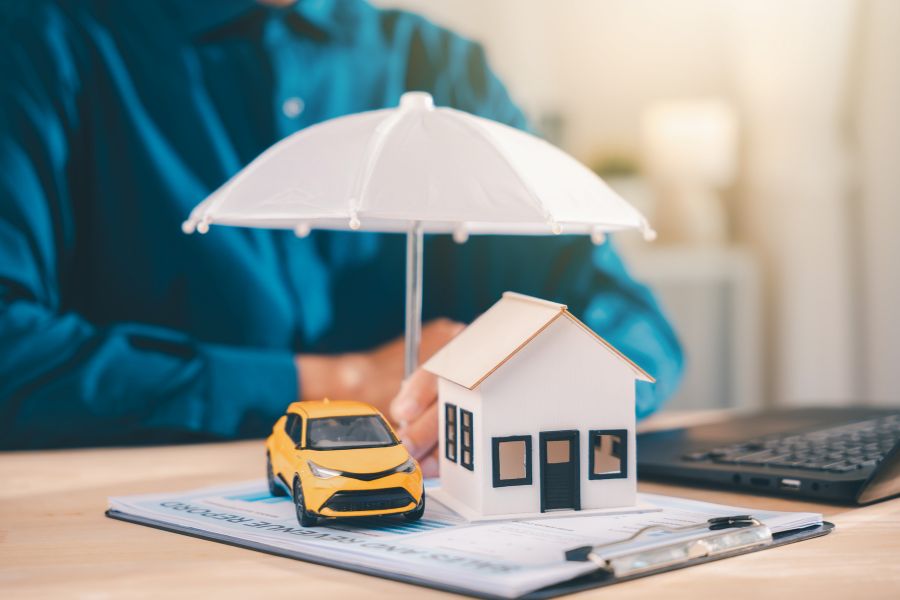 Une maquette de maison blanche et une voiture jaune sont protégées par un petit parapluie blanc ouvert sur des documents d'assurance.